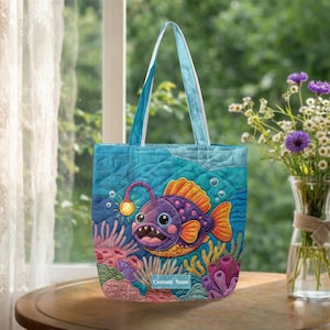 May include: A quilted tote bag featuring a colorful anglerfish design. The bag has a blue background with an underwater scene, including coral and bubbles. The anglerfish is purple, orange, and yellow. The text "Custom Name" is visible.