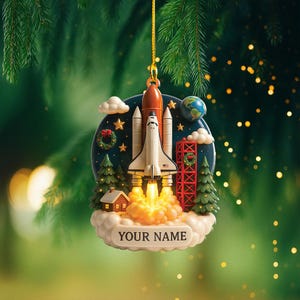 May include: A Christmas ornament featuring a space shuttle launching from a cloud base. The ornament includes a rocket, a globe, and a small house. The text "YOUR NAME" is displayed on the cloud base. The ornament is suspended by a gold string.