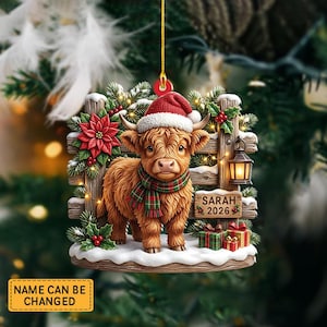 May include: A hanging Christmas ornament featuring a fluffy brown Highland cow wearing a red Santa hat and a green and red plaid scarf. The cow stands before a wooden fence with a lantern, poinsettias, and a sign reading SARAH 2026.