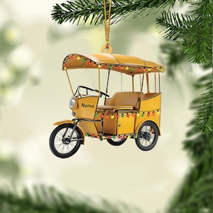 May include: Yellow tuk-tuk ornament with a canopy and string lights. The ornament has black wheels and a seat. The word "Name" is printed on the side. A gold string is attached for hanging. Festive holiday decor.