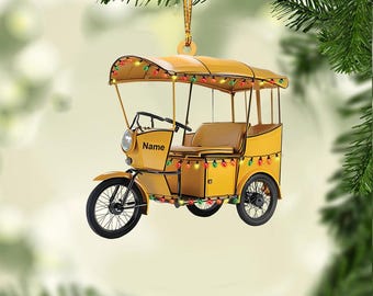 May include: Yellow tuk-tuk ornament with a canopy and string lights. The ornament has black wheels and a seat. The word "Name" is printed on the side. A gold string is attached for hanging. Festive holiday decor.