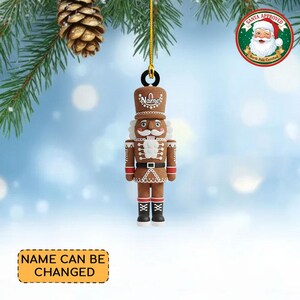 May include: A gingerbread-style nutcracker Christmas ornament with a brown body, red accents, and a white mustache. The ornament has a gold string for hanging and the text "Name" on the hat. The text "Name can be changed" is also visible.