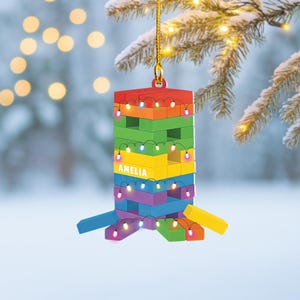 May include: A colorful Christmas ornament shaped like a tower of blocks, with the name "AMELIA" printed on it. The ornament is decorated with string lights and is suspended from a gold string. The blocks are red, orange, yellow, green, blue, and purple.