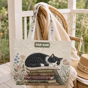 May include: Cream-colored tote bag featuring a sleeping black and white cat atop a stack of books. The bag has floral accents and the text "YOUR NAME" on a rectangular label. The handles are patterned with floral designs.