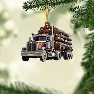 May include: A Christmas ornament shaped like a brown semi-truck hauling logs, decorated with colorful lights. The truck has the word "Name" on the front. The ornament is hanging from a gold string.