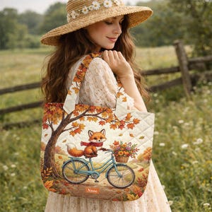 May include: A quilted tote bag featuring a printed illustration of a fox wearing a red scarf, riding a light blue bicycle. The scene includes a tree with orange autumn leaves and a basket of flowers. A rectangular label at the bottom reads Name.