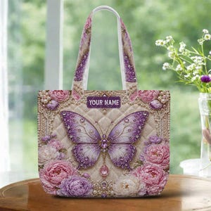 May include: A beige tote bag featuring a large purple butterfly with crystal accents and a pink teardrop pendant. The bag is adorned with pink and purple floral designs and the text "YOUR NAME". The handles and trim have a floral pattern.
