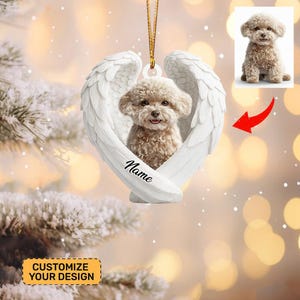 May include: A white heart-shaped ornament with angel wings cradles a small, fluffy dog. The dog has light brown fur and is smiling. The word "Name" is written on the ornament. A small image of the dog is in the upper right corner. The ornament hangs from a gold string.