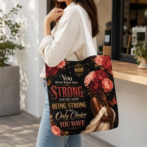 May include: Black tote bag with a floral design and the text "You never know how STRONG you are until BEING STRONG is the Only Choice YOU HAVE". The bag has a white strap and a floral design with red and pink flowers.