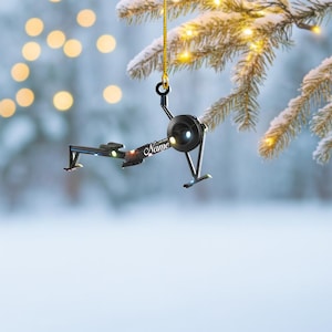 May include: A black and silver rowing machine ornament with the word "Name" in cursive. The ornament is hanging from a gold chain. The background is a blurred winter scene with lights.