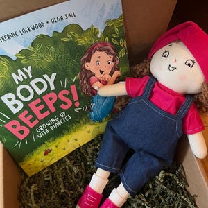 May include: A children's book titled "My Body Beeps! Growing Up With Diabetes" with a cartoon illustration of a girl on the cover. The book is in a cardboard box with green shredded paper and a handmade doll with a red shirt and blue overalls.