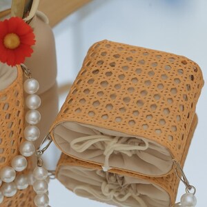 May include: A woven straw bag with a drawstring closure. The bag is light brown and has a woven pattern. The bag is sitting on a white surface.