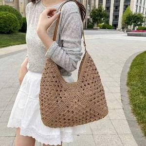 May include: A brown, open-weave shoulder bag with a brown leather strap. The bag has a large, curved shape and is made of a woven material. The bag is being held by a person wearing a grey long-sleeve shirt and a white skirt.