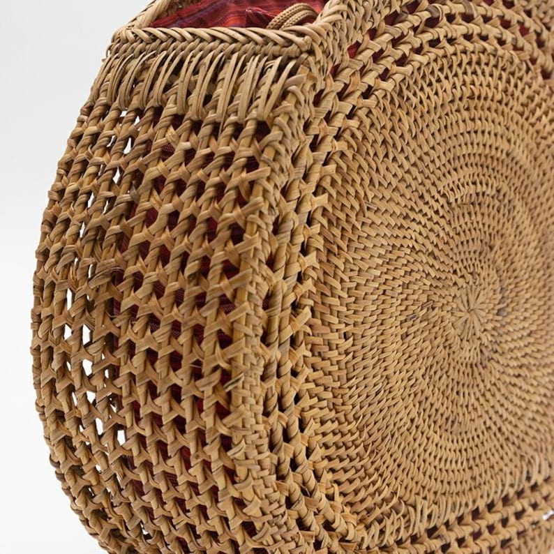 May include: A round, woven rattan bag with a spiral design. The bag is a light brown color and has a red fabric lining visible at the top. The weave is intricate and creates a textured surface.