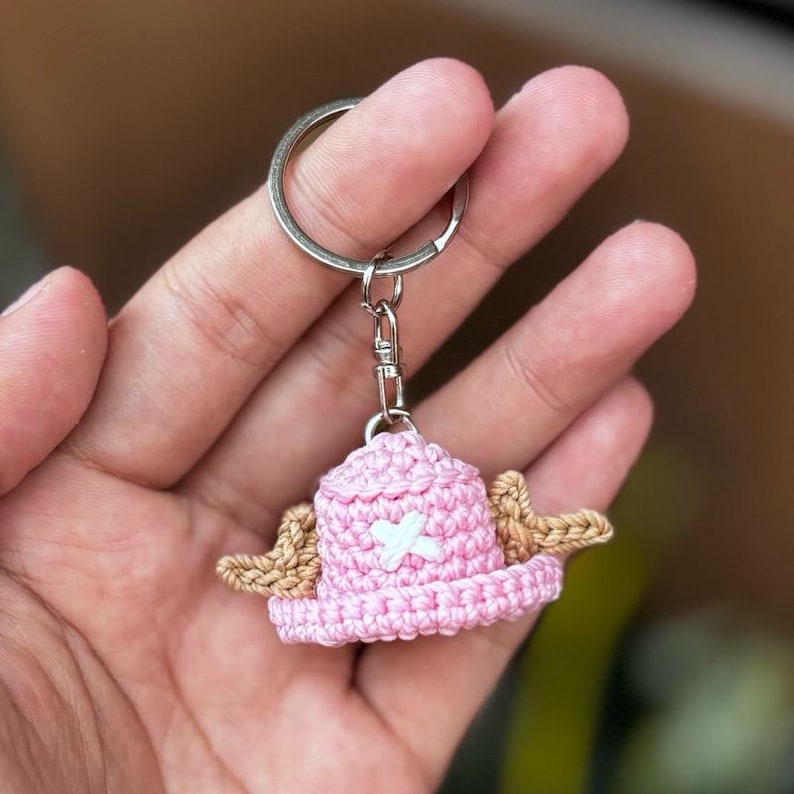 May include: A handmade pink crochet hat keychain with a white cross and tan wings. The keychain is attached to a silver metal ring and clasp. The hat is a whimsical accessory.