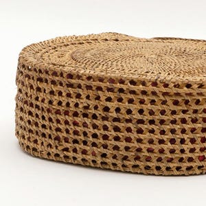 May include: A round, woven basket with a natural tan color. The basket has a textured, open weave design with a red interior visible through the openings. The top of the basket is also woven, and the overall shape is circular.