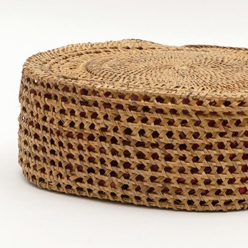 May include: A round, woven basket with a natural tan color. The basket has a textured, open weave design with a red interior visible through the openings. The top of the basket is also woven, and the overall shape is circular.