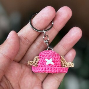 May include: A handmade pink crochet hat keychain with a white cross and tan accents. The keychain has a silver ring and clasp. The hat is a miniature version of a character's hat from a popular anime series.