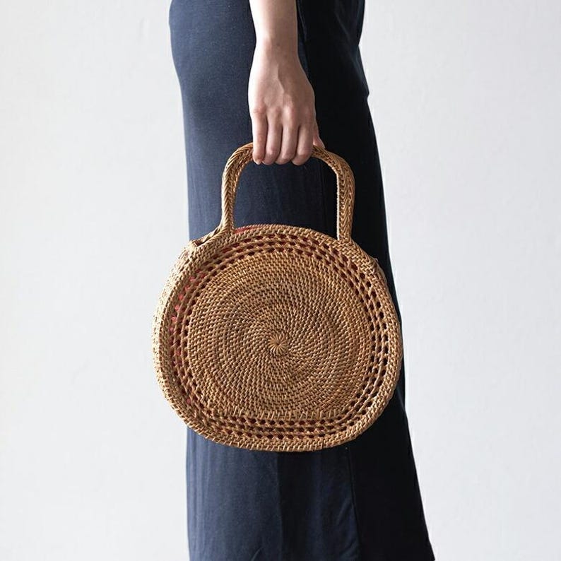 May include: A round, woven rattan handbag with two handles. The bag has a spiral design in the center and a reddish-brown interior. The bag is held by a person wearing a dark blue dress.