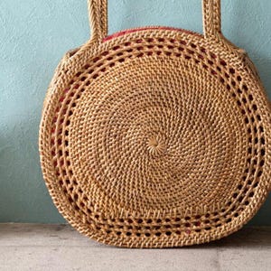 May include: A round, woven rattan handbag with two handles. The bag has a spiral design in the center and a red lining visible through the open weave. The bag is tan in color and sits on a gray surface.