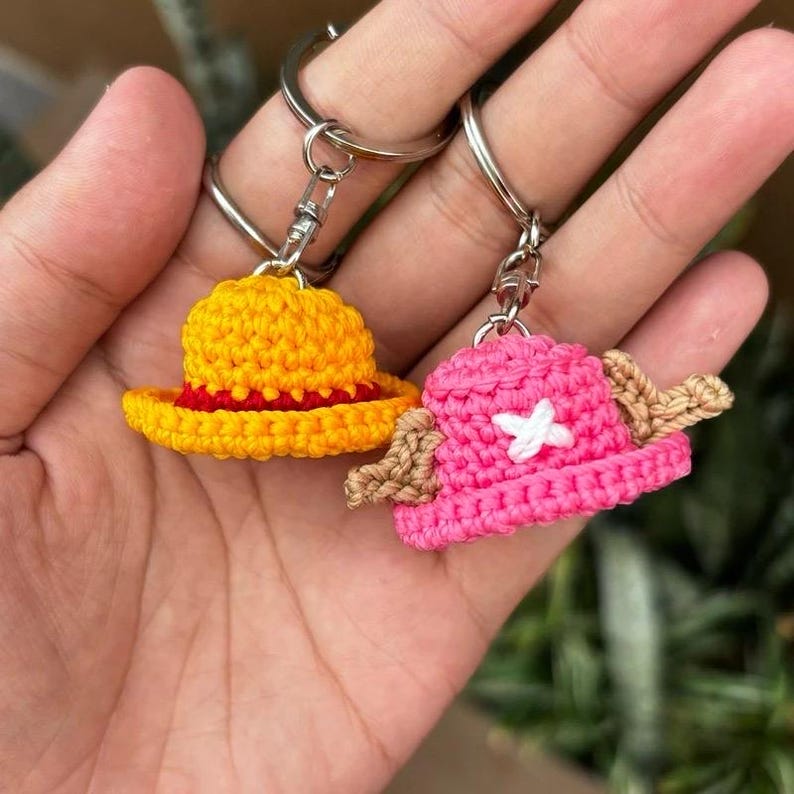 May include: Two crochet keychains featuring miniature hats. One hat is yellow with a red brim, the other is pink with a white cross and brown accents. Both keychains have silver-colored metal rings.