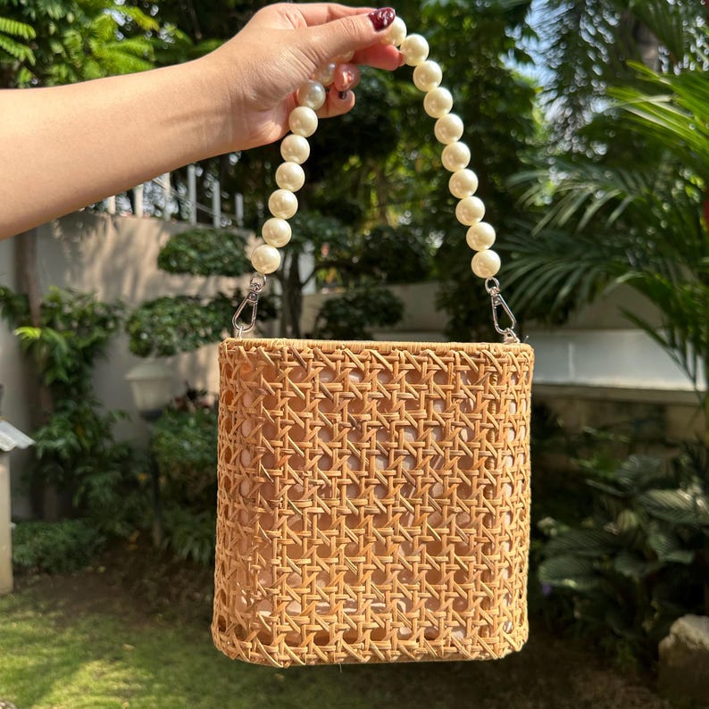 May include: A woven rattan bag with a square shape and a pearl handle. The bag has a light brown color and a woven pattern.