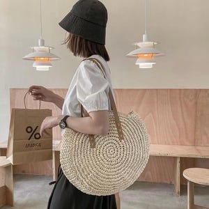 Pu&ograve; includere: Una grande borsa tote rotonda in paglia bianca con manici in pelle marrone. La borsa &egrave; grande e ha una trama intrecciata. La borsa &egrave; tenuta da una persona che indossa una camicia bianca e un cappello nero.