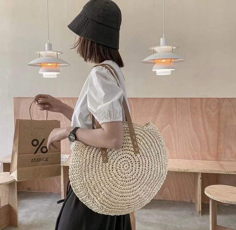 Pu&ograve; includere: Una grande borsa tote rotonda in paglia bianca con manici in pelle marrone. La borsa &egrave; grande e ha una trama intrecciata. La borsa &egrave; tenuta da una persona che indossa una camicia bianca e un cappello nero.