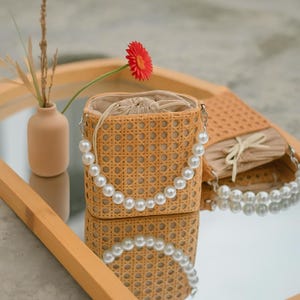 May include: A woven wicker bag with a pearl strap. The bag is light brown and has a woven pattern. The bag is sitting on a wooden surface with a mirror reflection.