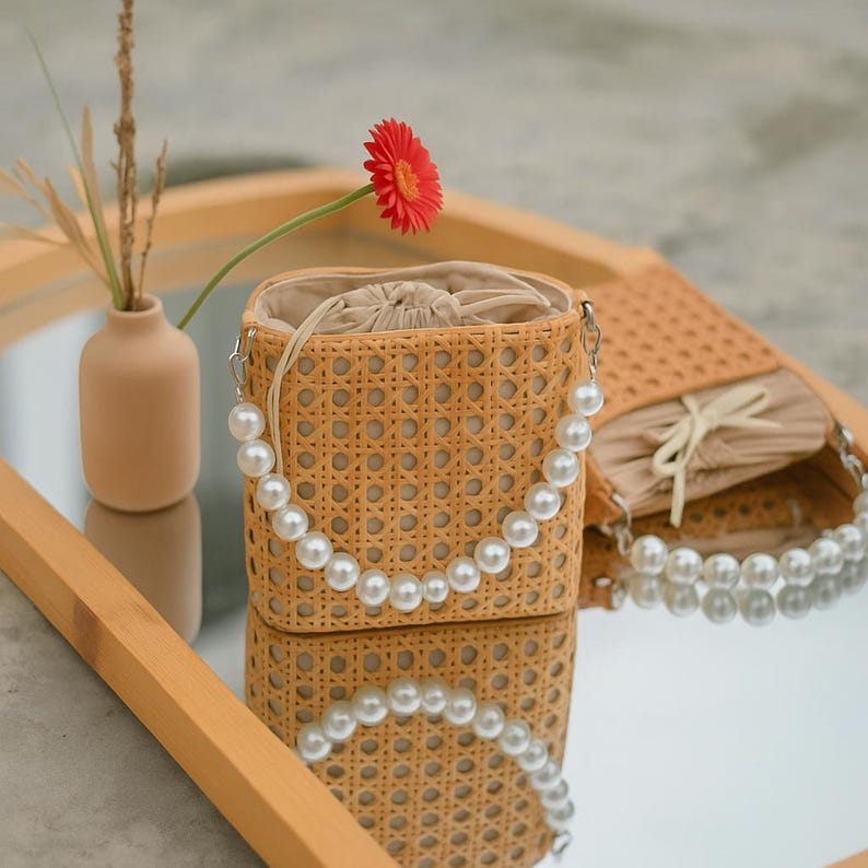 May include: A woven wicker bag with a pearl strap. The bag is light brown and has a woven pattern. The bag is sitting on a wooden surface with a mirror reflection.
