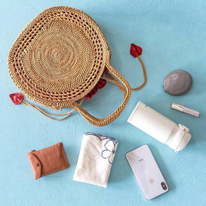 May include: A woven rattan handbag with a circular design, accompanied by a brown leather wallet, a white water bottle, a compact, a phone, a small towel with a floral design, and a tube of lip gloss. The items are arranged on a light blue surface.