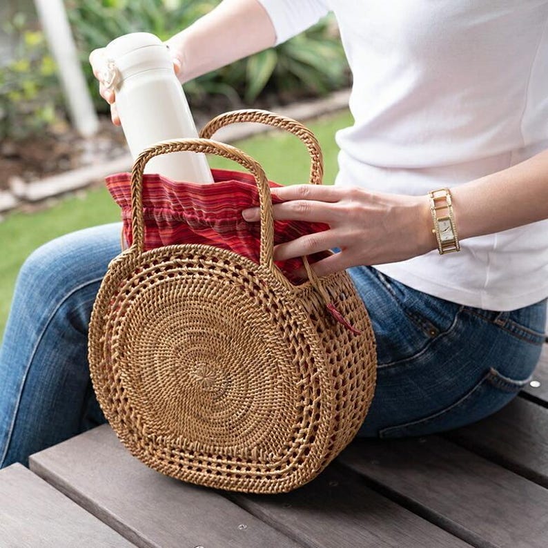 May include: A round, woven rattan handbag with two handles. The bag is open, revealing a red and white patterned fabric lining. A white water bottle is being placed inside the bag. The bag is tan in color.
