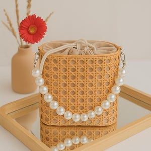 May include: A woven wicker bag with a pearl strap. The bag is light brown and has a white lining. The bag is sitting on a wooden tray with a mirror surface.