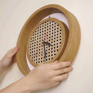 May include: A distinctive wall clock with a light brown wooden frame in a spiral design. The clock face has a woven rattan centre with black hands and a red second hand. The clock is held against a white wall.
