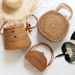 May include: An assortment of woven straw bags and accessories. The image features a round handbag with handles, a small half-moon bag, a bucket bag, a straw hat, and a black and white fan, all in natural tones.
