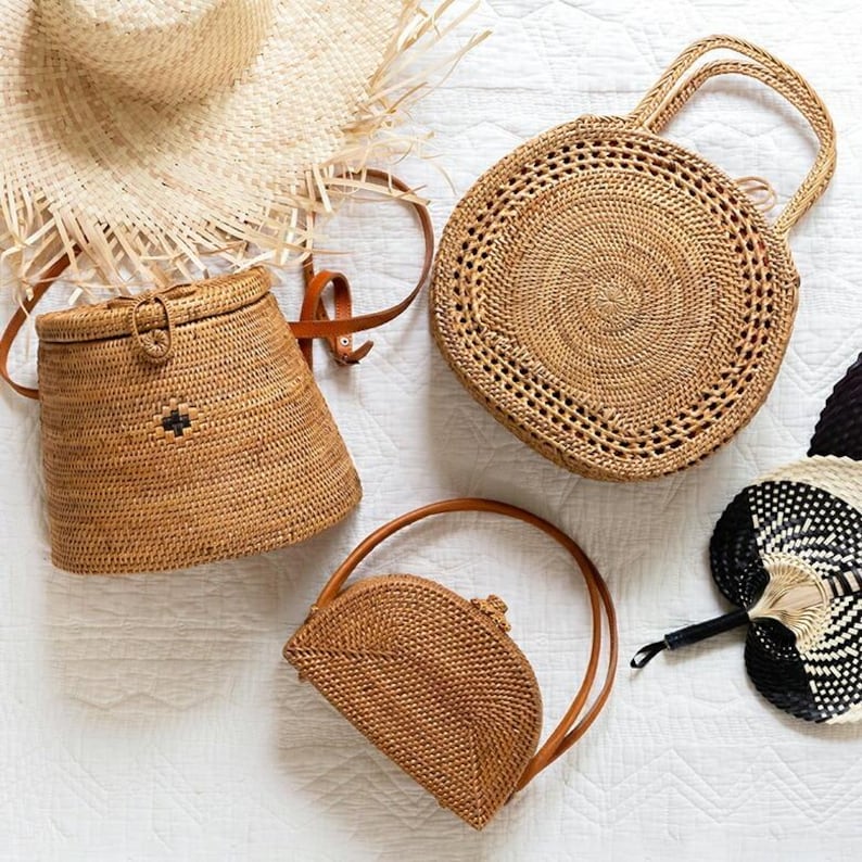 May include: An assortment of woven straw bags and accessories. The image features a round handbag with handles, a small half-moon bag, a bucket bag, a straw hat, and a black and white fan, all in natural tones.