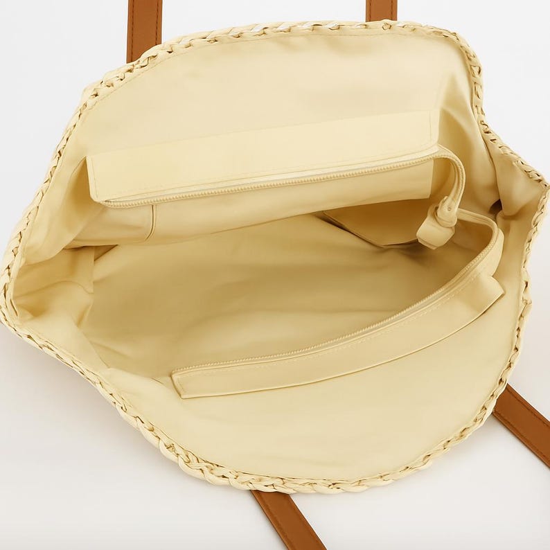 Pu&ograve; includere: Una borsa tote di paglia con manici in pelle marrone e una fodera in tessuto beige. La borsa ha una tasca con cerniera all'interno.