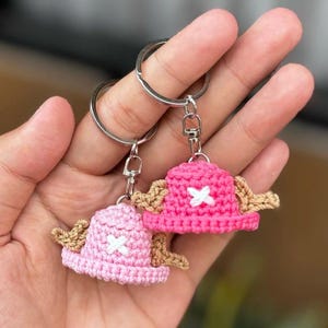 May include: Two crochet keychains featuring pink and light pink hats with white crosses and tan wings. The keychains are attached to silver metal rings and clasps. The hats are handmade and have a textured appearance.