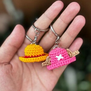 May include: Two crochet keychains featuring miniature hats. One is yellow with a red brim, the other is pink with a white cross and tan accents. Both are attached to silver keyrings.