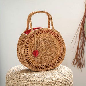 May include: A round, woven rattan bag with a red interior and a small red heart-shaped charm. The bag has two handles and is sitting on a woven stool. The bag is a natural tan color.