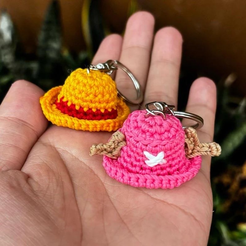 May include: Two crochet keychains shaped like hats. One is yellow with a red band, the other is pink with tan ear flaps and a white cross. Both have silver key rings attached.