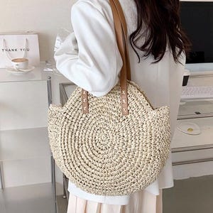 Pu&ograve; includere: Una borsa tote rotonda di paglia con manici in pelle marrone. La borsa ha un design intrecciato ed &egrave; di colore beige chiaro.