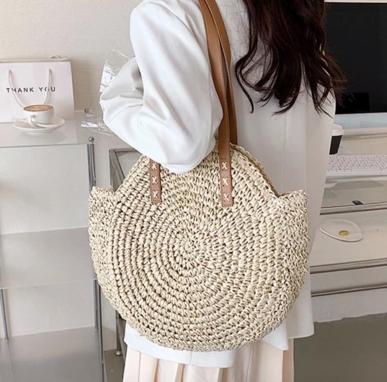 Pu&ograve; includere: Una borsa tote rotonda di paglia con manici in pelle marrone. La borsa ha un design intrecciato ed &egrave; di colore beige chiaro.