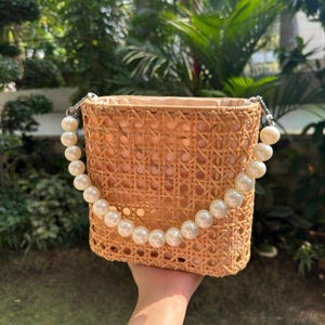 May include: A woven straw bag with a pearl handle. The bag is a light brown color and has a rectangular shape.