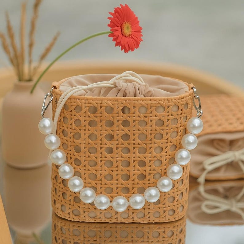 May include: A woven straw bag with a pearl strap. The bag is a light brown color and has a woven pattern. The strap is made of white pearls and is attached to the bag with silver hardware.