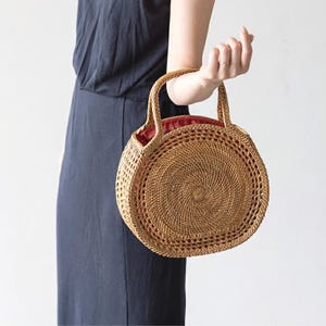 May include: A round, woven rattan handbag with two handles. The bag has a spiral design in the center and a red fabric lining. The bag is held by a person wearing a dark blue dress.