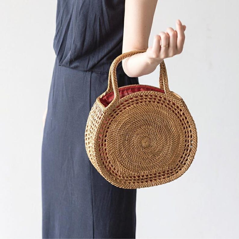 May include: A round, woven rattan handbag with two handles. The bag has a spiral design in the center and a red fabric lining. The bag is held by a person wearing a dark blue dress.
