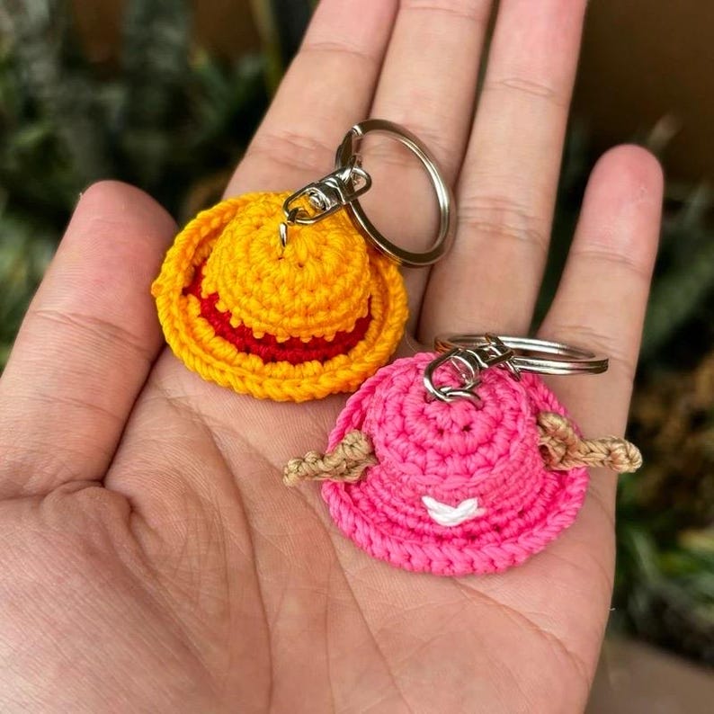 May include: Two crochet keychains shaped like hats. One is yellow with a red band, the other is pink with tan accents. Both have silver key rings attached. The hats are small and detailed, with a textured appearance.