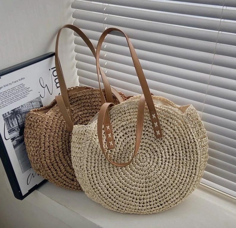Pu&ograve; includere: Due borse tote rotonde in paglia con manici in pelle marrone. La borsa a sinistra &egrave; marrone e la borsa a destra &egrave; beige.