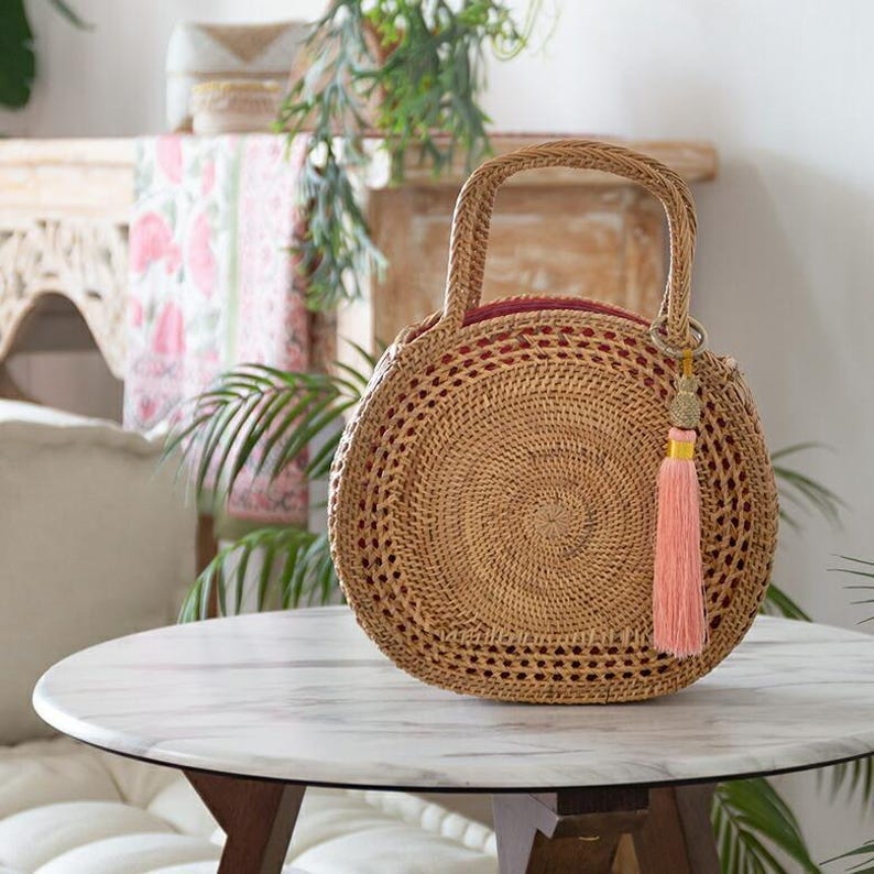 May include: Round woven rattan handbag with a handle and a pink tassel. The bag has a circular design with a woven pattern and a red interior. The bag is sitting on a white marble table.